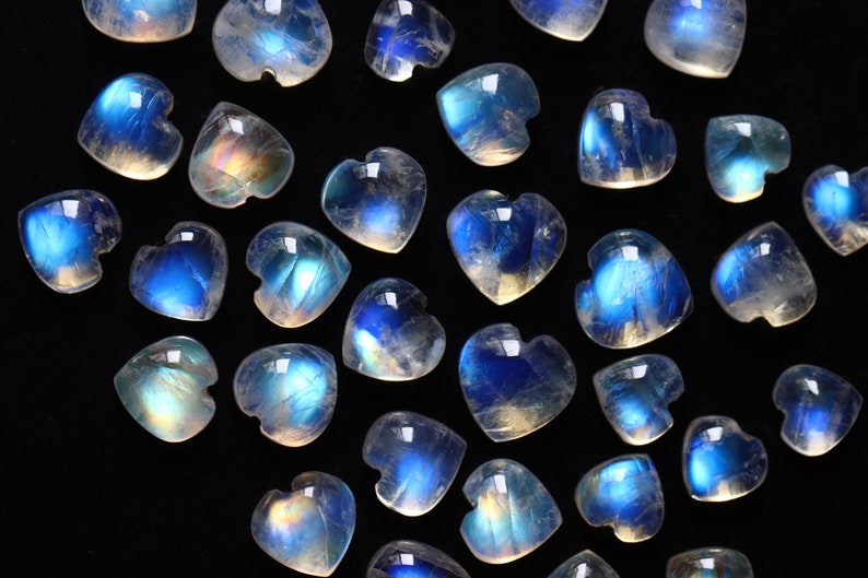 Puede incluir: Una colecci&oacute;n de peque&ntilde;os cabujones de piedra lunar en forma de coraz&oacute;n que presentan un aspecto transl&uacute;cido con una distintiva adularescencia azul. Cada piedra individual muestra una superficie lisa y pulida con una silueta de coraz&oacute;n redondeada.