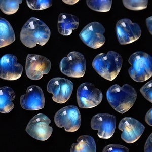 Puede incluir: Una colecci&oacute;n de peque&ntilde;os cabujones de piedra lunar en forma de coraz&oacute;n, con un aspecto transl&uacute;cido y lechoso con una distintiva adularescencia azul. Cada gema pulida muestra una superficie lisa y redondeada con un brillo iridiscente.