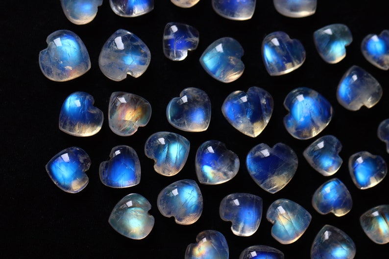 Puede incluir: Una colecci&oacute;n de peque&ntilde;os cabujones de piedra lunar en forma de coraz&oacute;n, con un aspecto transl&uacute;cido y lechoso con una distintiva adularescencia azul. Cada gema pulida muestra una superficie lisa y redondeada con un brillo iridiscente.