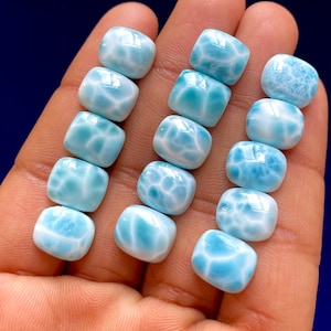 Puede incluir: Una colección de gemas de larimar pulidas, de corte cuadrado. Las piedras muestran un color azul pálido con patrones blancos en forma de telaraña. Las gemas se sostienen en una mano sobre un fondo azul oscuro.