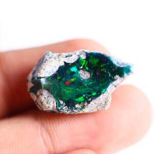 May include: A raw, unpolished opal gemstone featuring a deep, translucent green centre with vibrant flashes of emerald and red light, surrounded by a rough, textured, light grey rocky matrix that frames the colourful, crystalline interior.
