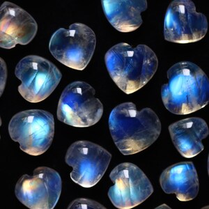 Puede incluir: Una colecci&oacute;n de peque&ntilde;os cabujones de piedra lunar en forma de coraz&oacute;n dispersos sobre un fondo negro. Cada piedra transl&uacute;cida muestra una adularescencia azul distintiva, reflejando la luz con un brillo iridiscente sobre las superficies.