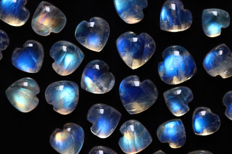 Puede incluir: Una colecci&oacute;n de peque&ntilde;os cabujones de piedra lunar en forma de coraz&oacute;n dispersos sobre un fondo negro. Cada piedra transl&uacute;cida muestra una adularescencia azul distintiva, reflejando la luz con un brillo iridiscente sobre las superficies.