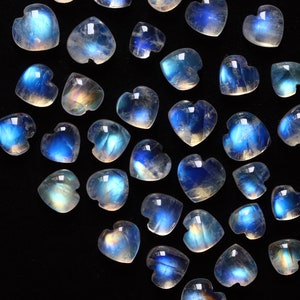 Puede incluir: Una colecci&oacute;n de peque&ntilde;os cabujones de piedra lunar en forma de coraz&oacute;n con una apariencia transl&uacute;cida y una distintiva adularescencia azul. Cada piedra preciosa muestra una superficie lisa y pulida con un brillo que cambia de intensidad.