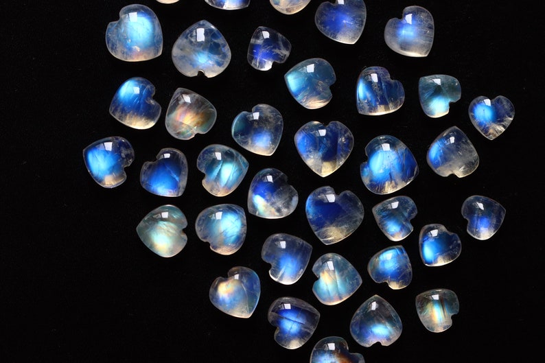 Puede incluir: Una colecci&oacute;n de peque&ntilde;os cabujones de piedra lunar en forma de coraz&oacute;n con una apariencia transl&uacute;cida y una distintiva adularescencia azul. Cada piedra preciosa muestra una superficie lisa y pulida con un brillo que cambia de intensidad.