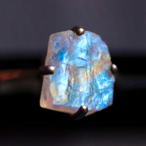 May include: A ring featuring a raw, translucent gemstone with a prominent blue iridescent sheen. The stone has an irregular, jagged shape and is secured by four dark metal prongs that extend from a thin, dark metal band, creating a contrast.