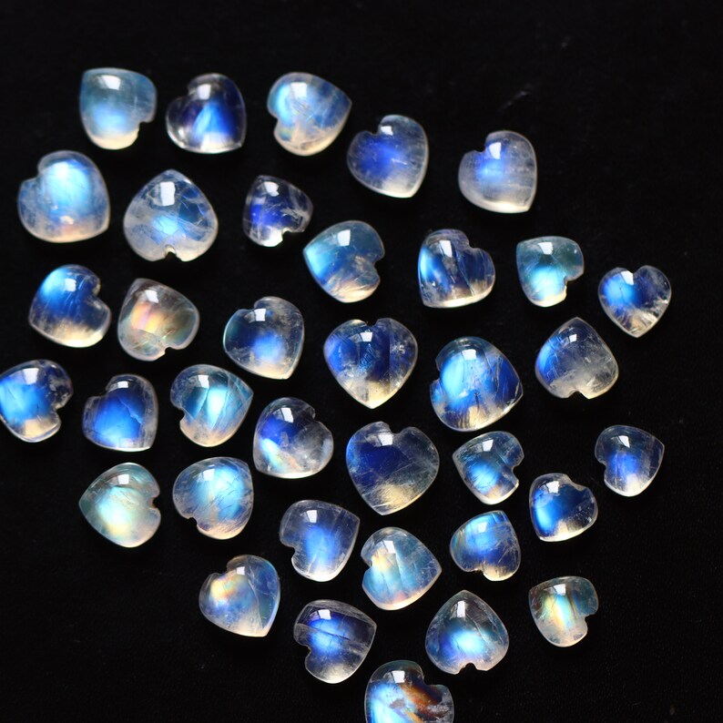 Puede incluir: Una colecci&oacute;n de peque&ntilde;os cabujones de piedra lunar en forma de coraz&oacute;n, con un aspecto transl&uacute;cido y lechoso con destellos azules iridiscentes. Cada gema individual muestra una superficie pulida y una silueta de coraz&oacute;n.