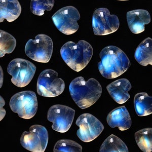 Puede incluir: Una colecci&oacute;n de corazones de piedra lunar pulida, cada uno con una apariencia transl&uacute;cida y lechosa con una distintiva adularescencia o destello azul. Las piedras preciosas en forma de coraz&oacute;n est&aacute;n dispuestas en un patr&oacute;n disperso.
