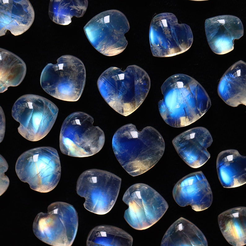 Puede incluir: Una colecci&oacute;n de corazones de piedra lunar pulida, cada uno con una apariencia transl&uacute;cida y lechosa con una distintiva adularescencia o destello azul. Las piedras preciosas en forma de coraz&oacute;n est&aacute;n dispuestas en un patr&oacute;n disperso.