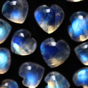 Puede incluir: Una colecci&oacute;n de peque&ntilde;os cabujones de piedra lunar en forma de coraz&oacute;n con una apariencia blanca lechosa y transl&uacute;cida con destellos azules iridiscentes. Cada piedra muestra una superficie lisa y pulida con una silueta de coraz&oacute;n definida.
