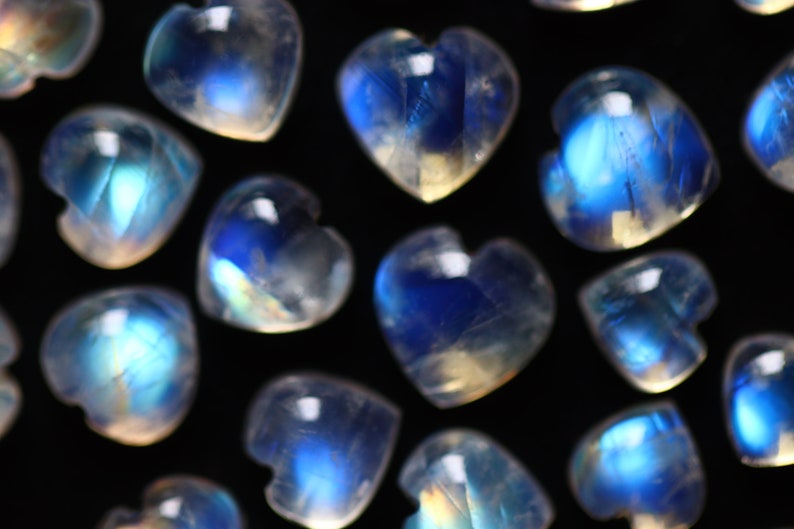 Puede incluir: Una colecci&oacute;n de peque&ntilde;os cabujones de piedra lunar en forma de coraz&oacute;n con una apariencia blanca lechosa y transl&uacute;cida con destellos azules iridiscentes. Cada piedra muestra una superficie lisa y pulida con una silueta de coraz&oacute;n definida.
