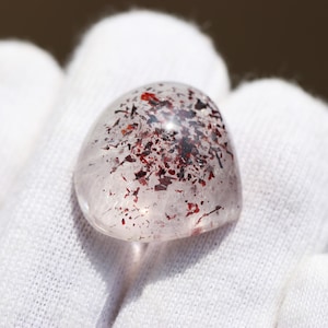 Peut inclure: Une pierre précieuse polie, translucide et arrondie, présentant des inclusions internes de flocons minéraux rouge foncé et noirs dispersés dans la structure de quartz clair, créant un motif tacheté distinct sur la surface lisse en dôme.