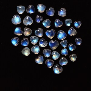 Puede incluir: Una colecci&oacute;n de peque&ntilde;os cabujones de piedra lunar en forma de coraz&oacute;n dispuestos en un grupo. Cada piedra preciosa muestra una apariencia transl&uacute;cida y lechosa con una distintiva adularescencia azul o destello de luz sobre la superficie pulida.