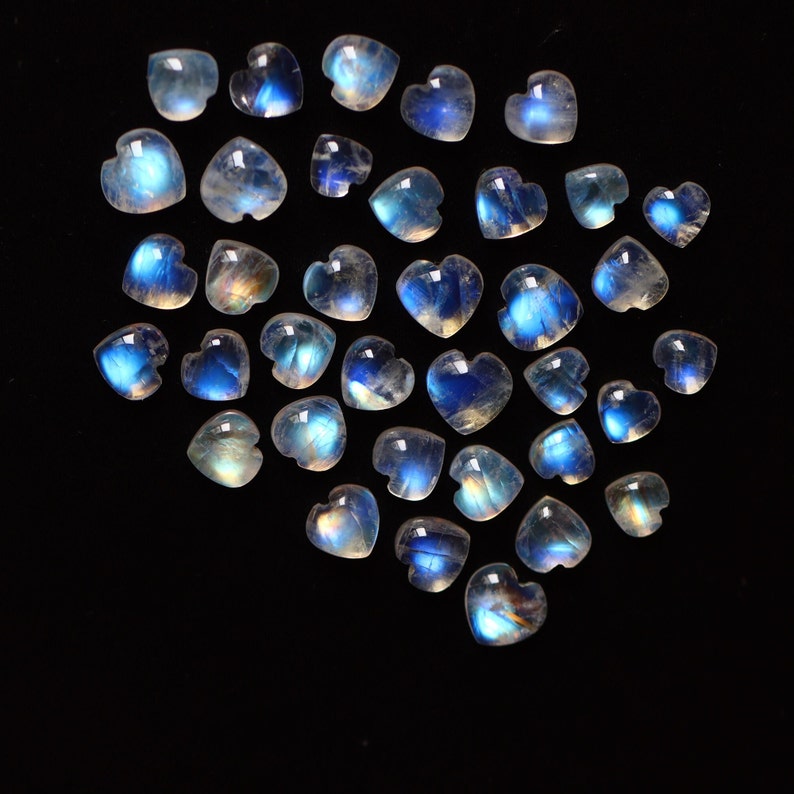Puede incluir: Una colecci&oacute;n de peque&ntilde;os cabujones de piedra lunar en forma de coraz&oacute;n dispuestos en un grupo. Cada piedra preciosa muestra una apariencia transl&uacute;cida y lechosa con una distintiva adularescencia azul o destello de luz sobre la superficie pulida.