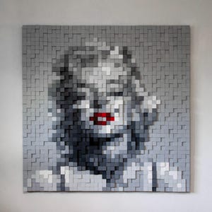 Puede incluir: Una pieza de arte mural cuadrada compuesta por numerosos bloques cuadrados pequeños y elevados en varios tonos de gris, blanco y negro, formando un retrato pixelado de una persona con labios de color rojo brillante como único acento.
