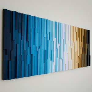 Puede incluir: Una pieza de arte de pared hecha de listones de madera en tonos de azul, blanco, beige, marrón y negro. Los listones están dispuestos en un patrón de degradado, creando un diseño moderno y abstracto.