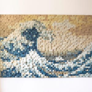 Puede incluir: Una pieza de arte de pared que representa la Gran Ola de Kanagawa, una famosa estampa japonesa en madera de Hokusai, hecha de pequeños bloques de madera en tonos azules, blancos y beige.