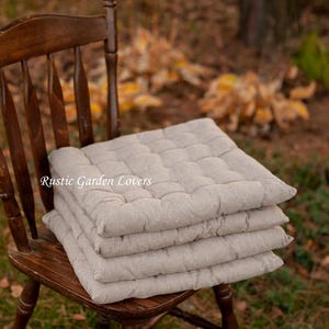 Puede incluir: Una pila de cuatro cojines de asiento cuadrados y acolchados de color beige sobre una silla de madera de color marrón oscuro con respaldo de husillos. El texto "Rustic Garden Lovers" está impreso en blanco en el centro de los cojines.