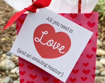 May include: A pink gift bag with a white tag that says "All you need is Love (and an amazing Realtor)". The tag has a red heart with the word "Love" in white. The bag is decorated with pink hearts.