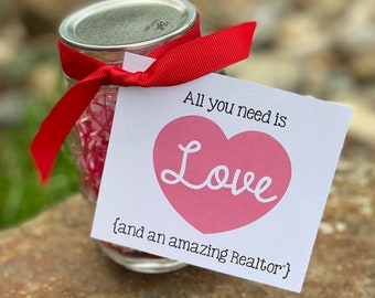 May include: A white card with red heart and the text "All you need is Love {and an amazing Realtor}" tied with a red ribbon to a clear glass jar filled with pink and white confetti.