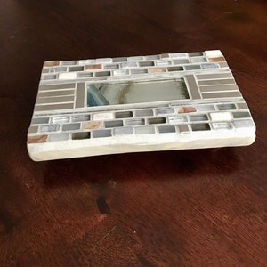 Op de afbeelding: Een rechthoekige mozaïekspiegel met een witte achtergrond en een verscheidenheid aan grijze, bruine en witte tegels. De spiegel is omlijst met een grijze rand en heeft een kleine, rechthoekige spiegel erin.