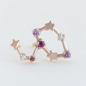 Op de afbeelding: Gouden sterrenbeeld oorbel met roze, paarse en witte edelstenen. Het sterrenbeeld heeft de vorm van een ster met een gebogen staart.
