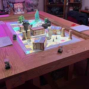 May include: A wooden tabletop game board with a light pink glow, featuring miniature buildings and figures. The board is surrounded by a dark wood frame and has a white surface with a printed map. The board is set up for a tabletop role-playing game.