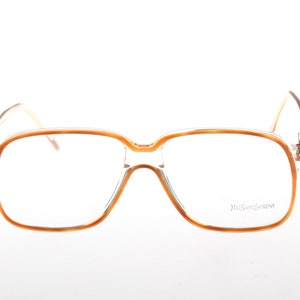 Może przedstawiać: Okulary w stylu vintage z kwadratową oprawką i jasnobrązową obwódką. Soczewki są przezroczyste, a zauszniki są ciemnobrązowe. Nazwa marki "Yves Saint Laurent" jest wydrukowana na prawej soczewce.