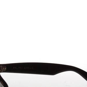 Peut inclure: Gros plan sur la branche en plastique noire d'une paire de lunettes de soleil. La branche est grav&eacute;e du texte "B&L RAY-BAN U.S."