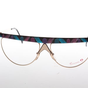 Puede incluir: Un par de gafas con montura dorada y un marco estampado en negro y azul. Las gafas tienen un diseño geométrico y el nombre de la marca "Casanova" está impreso en la patilla derecha.