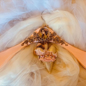 Könnte beinhalten: Ein dekorativer, pfirsichfarbener Holzkleiderbügel mit einem goldenen Metallhaken. Der Kleiderbügel zeigt ein Porträt einer Frau und aufwendige goldene Blumendetails. Ein gold-rosa herzförmiges Ornament mit Perlenbesatz hängt in der Mitte.