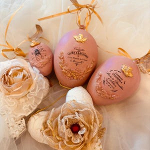 Op de afbeelding: Een verzameling paasdecoraties, waaronder roze eieren met gouden details en Franse tekst. Andere ornamenten zijn voorzien van een wit en goud bloemmotief en een kanten bloem, allemaal met een vintage esthetiek.