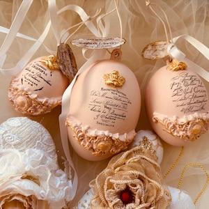 Può includere: Tre ornamenti decorativi a forma di uovo, in rosa tenue, adornati con dettagli dorati e ornati. Ogni uovo presenta una scrittura elegante ed è sospeso da un nastro bianco. Gli ornamenti hanno un'estetica vintage e antica.