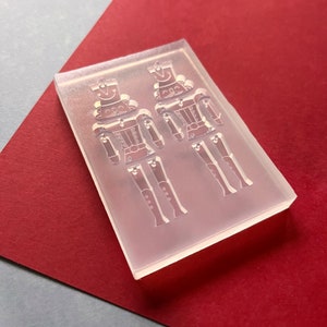 May include: A clear, rectangular mould featuring two soldier designs. The soldier figures have hats, jackets, and articulated legs. The mould is set against a red and grey background, suggesting a craft or DIY project.
