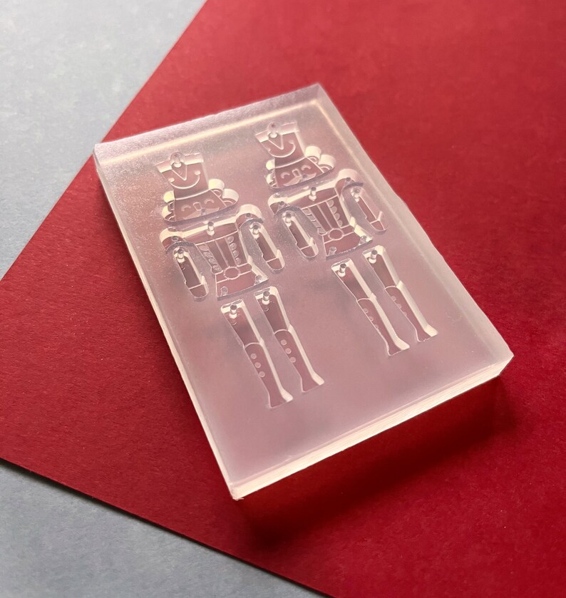 May include: A clear, rectangular mould featuring two soldier designs. The soldier figures have hats, jackets, and articulated legs. The mould is set against a red and grey background, suggesting a craft or DIY project.