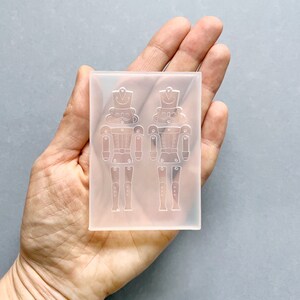 May include: A clear silicone mould in a rectangular shape, featuring two detailed nutcracker soldier designs. The mould is held in a hand, highlighting the intricate details of the toy soldier figures, including their hats, uniforms, and articulated limbs. The mould is for crafting.