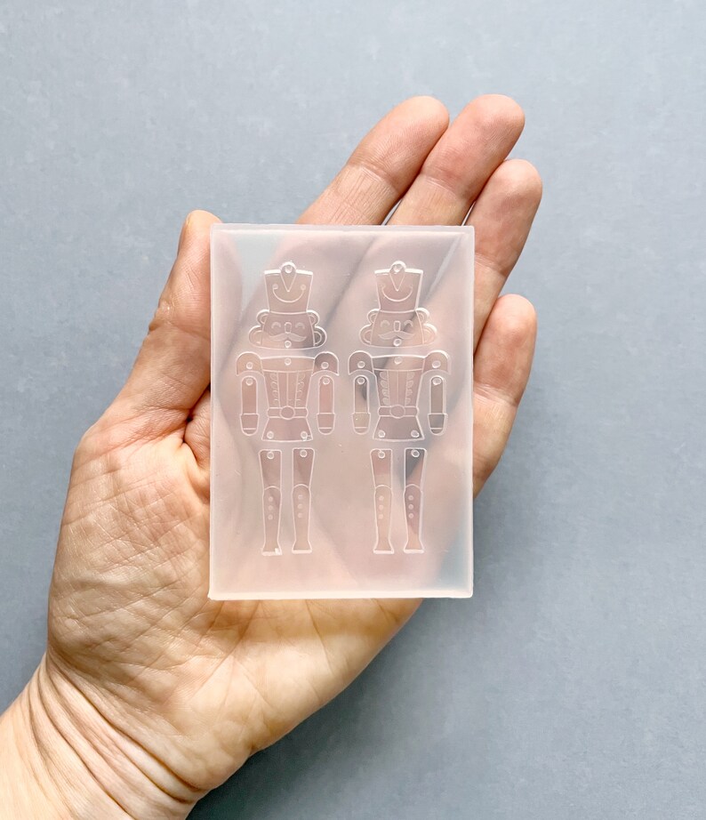May include: A clear silicone mould in a rectangular shape, featuring two detailed nutcracker soldier designs. The mould is held in a hand, highlighting the intricate details of the toy soldier figures, including their hats, uniforms, and articulated limbs. The mould is for crafting.