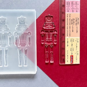May include: A clear resin mould and a finished resin nutcracker figurine. The mould features two nutcracker shapes, while the figurine stands next to a ruler, approximately 15 cm tall, on a red and grey surface.