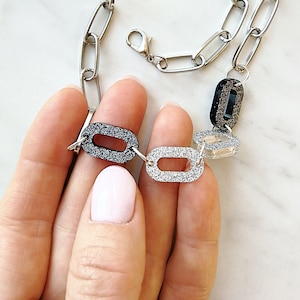 Puede incluir: Un collar de cadena plateada con dijes ovalados negros, plateados y transparentes. Los dijes est&aacute;n cubiertos de purpurina.