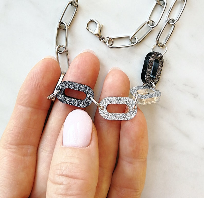 Puede incluir: Un collar de cadena plateada con dijes ovalados negros, plateados y transparentes. Los dijes est&aacute;n cubiertos de purpurina.