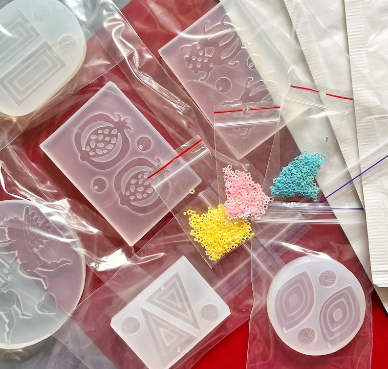 May include: A selection of clear silicone moulds in various shapes, including geometric designs, a pomegranate, and a mythical creature. Small bags of colourful star-shaped beads are also visible, along with other craft supplies.