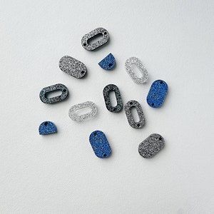 Puede incluir: Surtido de adornos ovalados cubiertos de purpurina en negro, plateado y azul. Los adornos tienen un agujero en el centro para enhebrar.