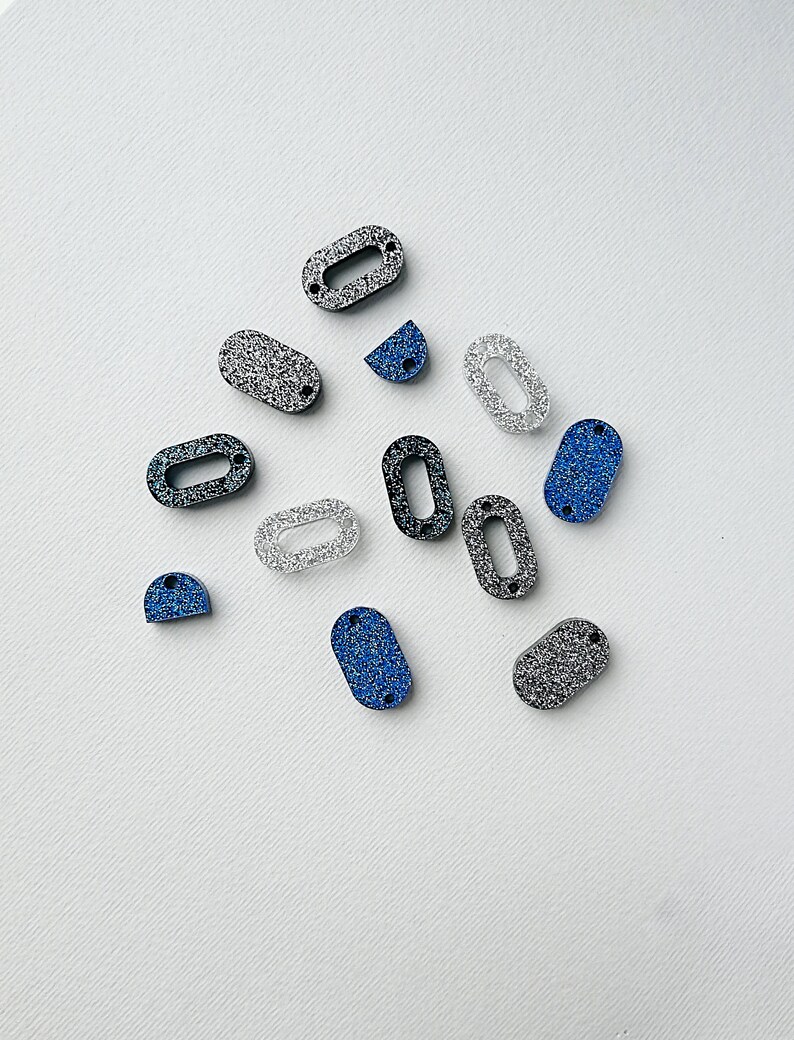 Puede incluir: Surtido de adornos ovalados cubiertos de purpurina en negro, plateado y azul. Los adornos tienen un agujero en el centro para enhebrar.