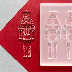 May include: A clear acrylic nutcracker charm and a silicone mould with nutcracker impressions. The charm is on a red background with a diagonal grey section. The nutcracker has a hat, moustache, and uniform.