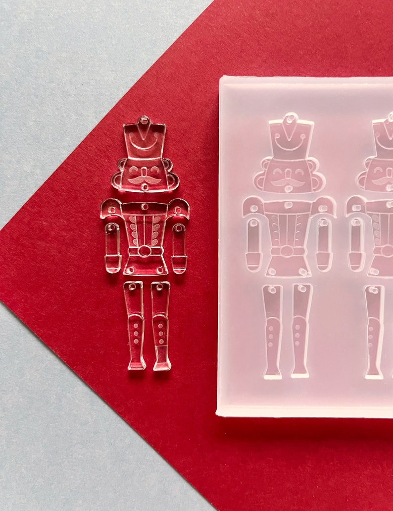 May include: A clear acrylic nutcracker charm and a silicone mould with nutcracker impressions. The charm is on a red background with a diagonal grey section. The nutcracker has a hat, moustache, and uniform.