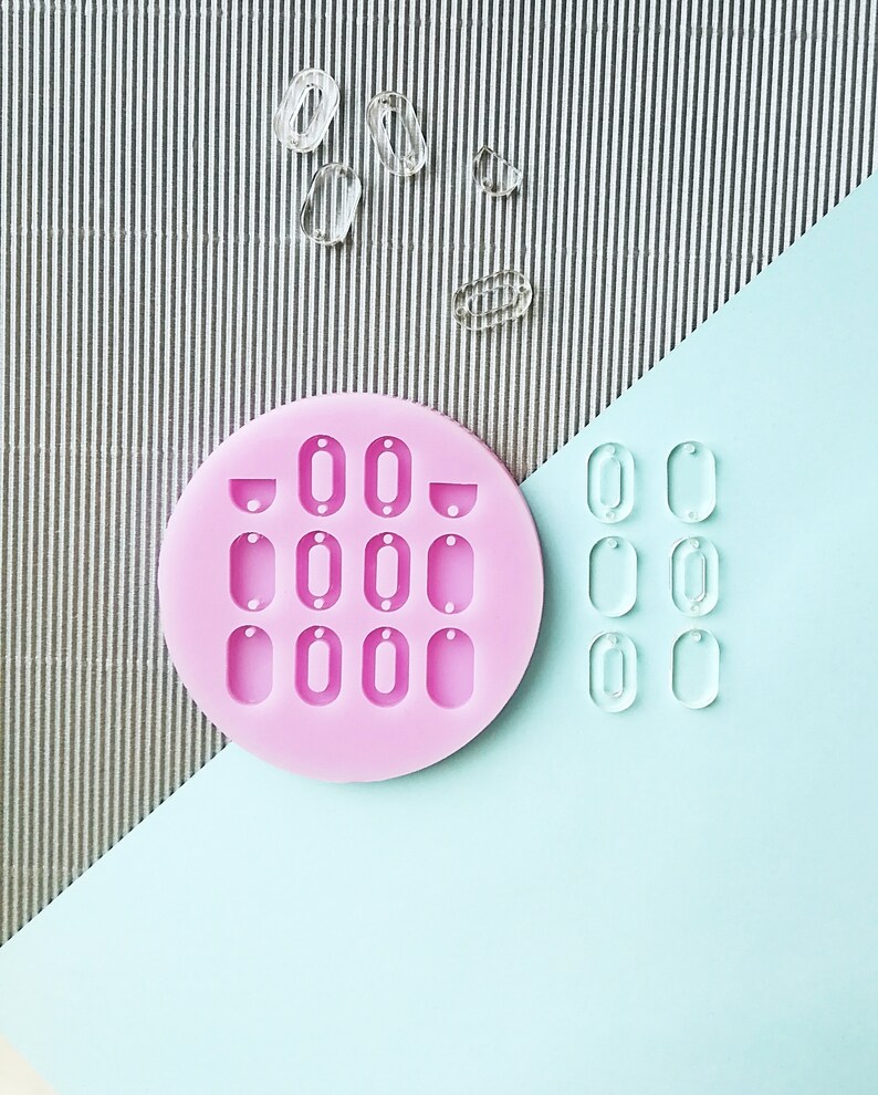 Puede incluir: Molde de silicona rosa con 12 cavidades ovaladas para hacer pendientes de resina. El molde est&aacute; sobre una superficie azul claro con pendientes de resina transparente.