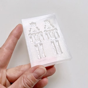 May include: A clear, rectangular mould featuring two detailed nutcracker soldier designs. The mould is held in a hand against a white background. The nutcrackers have hats, moustaches, and buttoned jackets, ideal for festive crafts.