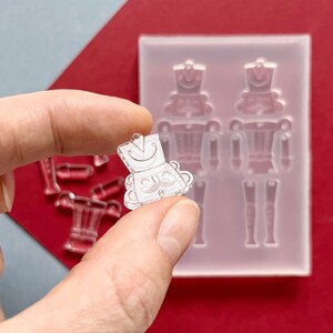 May include: A clear, miniature nutcracker charm held between fingers, with a matching mold in the background. The charm has a smiling face, a hat, and a mustache. Additional clear resin pieces are visible.