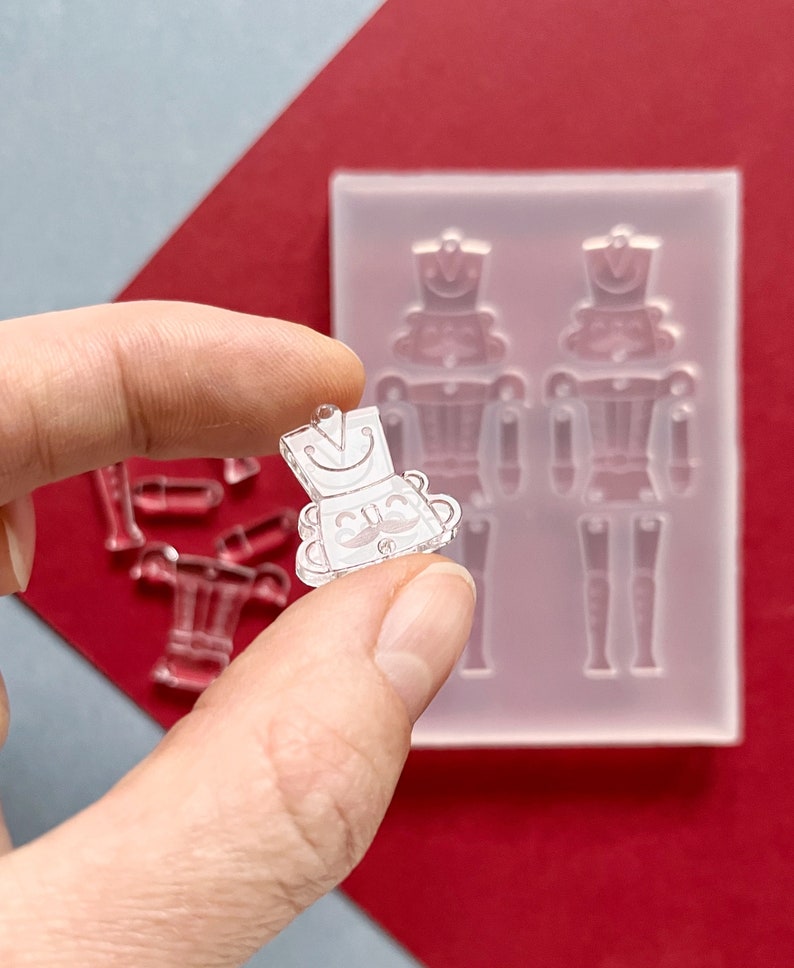 May include: A clear, miniature nutcracker charm held between fingers, with a matching mold in the background. The charm has a smiling face, a hat, and a mustache. Additional clear resin pieces are visible.