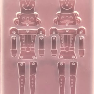 May include: A clear, flexible mould featuring two detailed nutcracker soldier designs. Each soldier has a hat, moustache, and uniform, with separate leg pieces. The mould is designed for crafting decorative items.